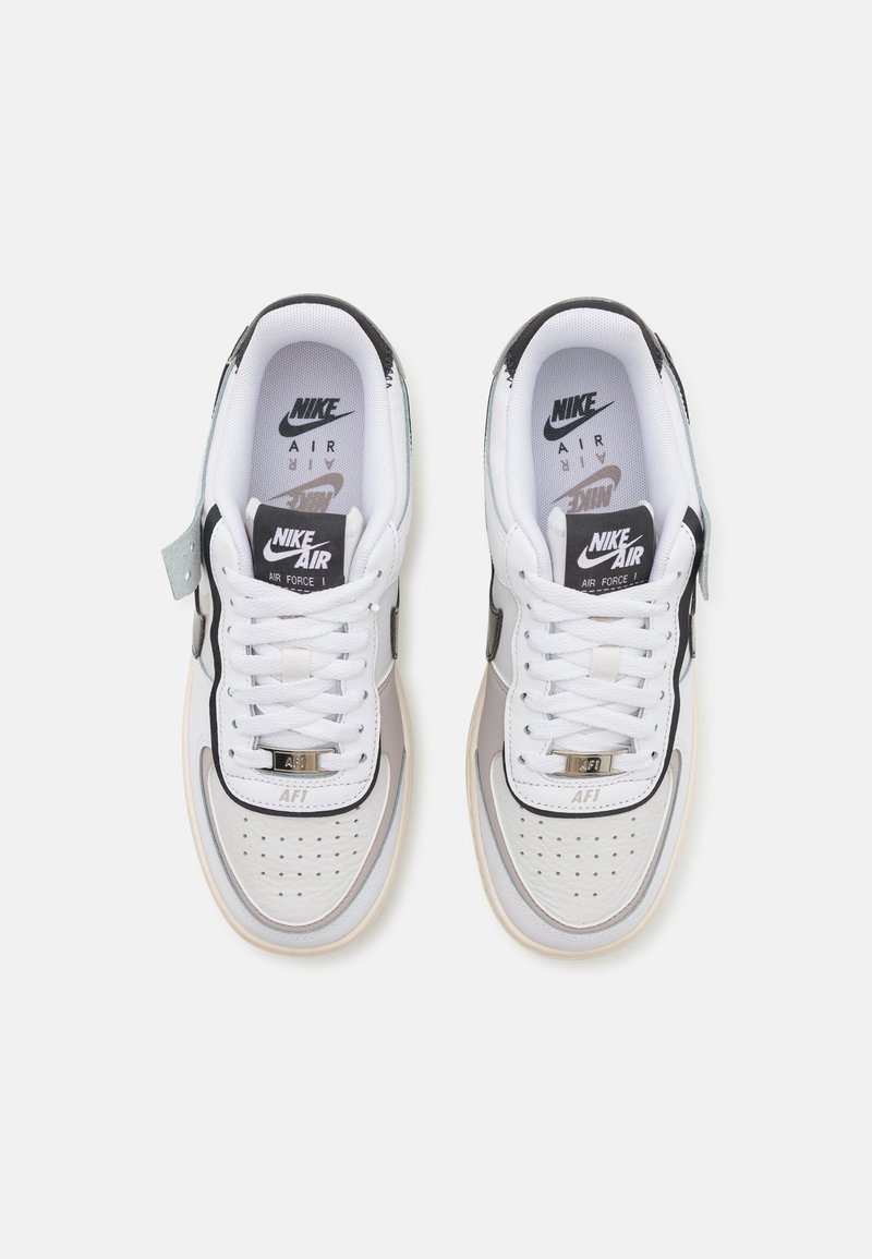 Zapatillas Nike Air Force 1 blancas con acentos negros, etiqueta en la lengüeta gris y puntera perforada. Presentan cordones blancos y una plantilla de marca visible.