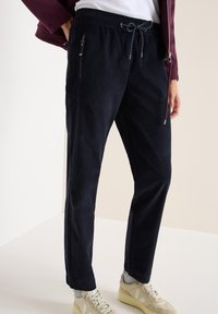 Granatowe spodnie ze sznurkiem w pasie z zamkami błyskawicznymi, wykonane z miękkiego materiału; mają luźny krój i zwężane nogawki, w połączeniu z jasnymi sneakersami.