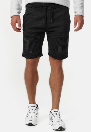 Mand iført sorte ribbede og distressed shorts med snøre i taljen og hvide New Balance-sneakers mod hvid baggrund.