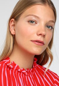 Blusa a rayas rojas y rosas con escote con volantes. La modelo, con cabello largo y rubio, lleva pendientes de aro pequeños y un maquillaje natural.