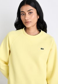 Sweatshirt amarelo com decote redondo e mangas raglan. Apresenta um logo verde da Lacoste no peito esquerdo. Textura de tecido suave.