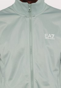 Jachetă verde deschis, cu fermoar, fabricată dintr-un material neted. Are guler înalt, o ușoară strălucire, logo imprimat pe pieptul stâng și un fermoar complet.