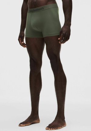 Hombre con calzoncillos bóxer Lululemon verde oliva, de pie descalzo frente a un fondo claro y liso.