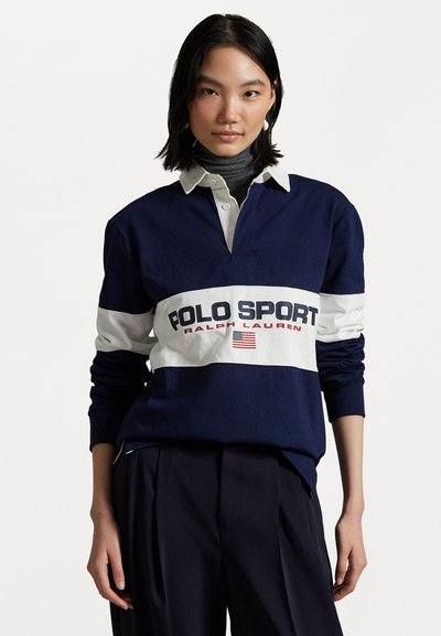 Jūras zila regbija stila krekls ar baltu horizontālu svītru, kurā ir teksts "POLO SPORT" un "RALPH LAUREN", garām piedurknēm un ar apkakli.