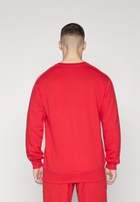 Uomo con capelli corti che indossa una felpa rossa a maniche lunghe e pantaloni rossi abbinati, visto da dietro su uno sfondo semplice.