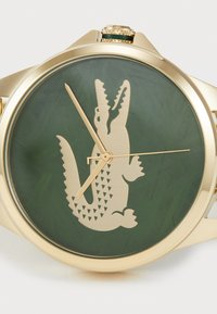 Goldfarbene Uhr mit einem grünen strukturierten Zifferblatt, das eine große Krokodilsilhouette zeigt, goldenen Zeigern und einem gemusterten Sekundenzeiger.