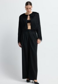 Kobieta ubrana w czarny, fakturowany krótki kardigan na czarnym biustonoszu, szerokie czarne spodnie oraz czarne szpilki, stojąca na tle białym.