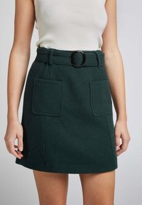 Jupe en mélange de laine vert foncé avec taille haute, large passant de ceinture, boucle ronde noire et deux poches plaquées à l'avant.