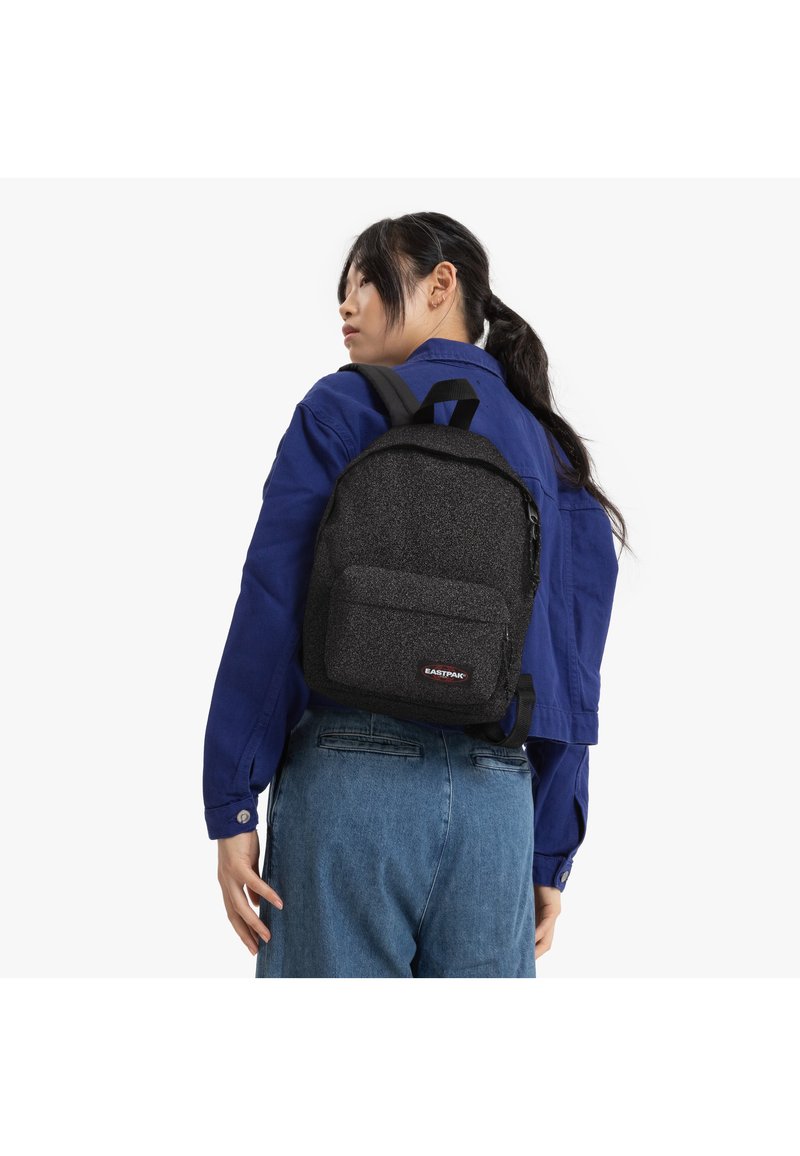 Un sac à dos noir texturé avec une poche avant, des straps ajustables et une poignée supérieure, porté sur le dos d'une personne portant une chemise bleue.
