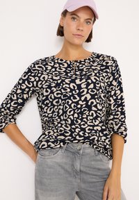 Maglietta a maniche lunghe blu navy con stampa leopardata beige, caratterizzata da un collo rotondo e dettaglio a chiave. Abbinata a pantaloni in denim grigio chiaro.