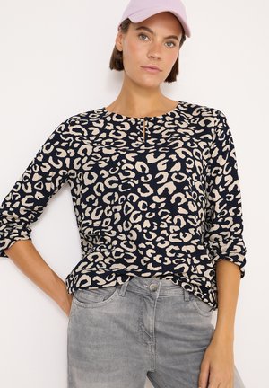 Maglietta a maniche lunghe blu navy con stampa leopardata beige, caratterizzata da un collo rotondo e dettaglio a chiave. Abbinata a pantaloni in denim grigio chiaro.