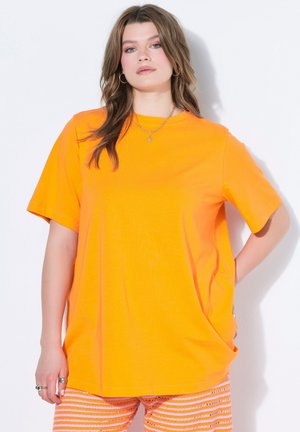 Vrouw met golvend haar draagt een grote oranje t-shirt en gestreepte roze en oranje broek, staand tegen een witte achtergrond met hand in de zak.