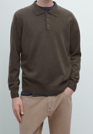 Homme portant un pull polo marron à manches longues sur une chemise sombre avec un pantalon beige, une main dans la poche, devant un fond clair uni.
