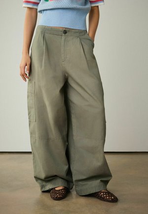 Person wearing wide-leg olive green cargo pants and brown woven slip-on shoes standing with one hand in pocket.