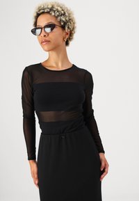 Schwarzes Langarmoberteil mit durchsichtigen Einsätzen, kombiniert mit einem schwarzen Kleid mit Kordelzug-Taille. Eine Cat-Eye-Sonnenbrille rundet den Look ab.