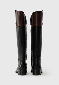 Bottes hauteur genou en cuir noir avec un accent en cuir marron en haut, des lacets à l'arrière et un petit talon carré. Design robuste et élégant.