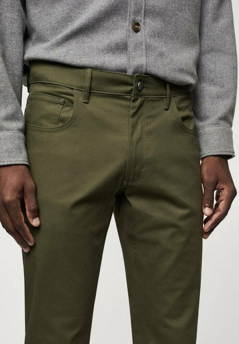 Olivgrüne, schmal geschnittene Hose aus weichem Stoff. Mit seitlichen Taschen und einem vorderen Knopfverschluss, kombiniert mit einem grauen Hemd mit Knöpfen.