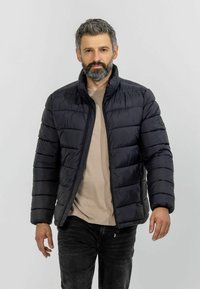 Black padded jacket with a high collar, horizontal quilting, and zipper pockets, paired with a beige shirt and dark jeans.