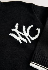 Černý pletený svetr s bílým logem a texturovanými akcenty. Žebrované manžety s bílými pruhy zdůrazňují design. Výstřih do V.