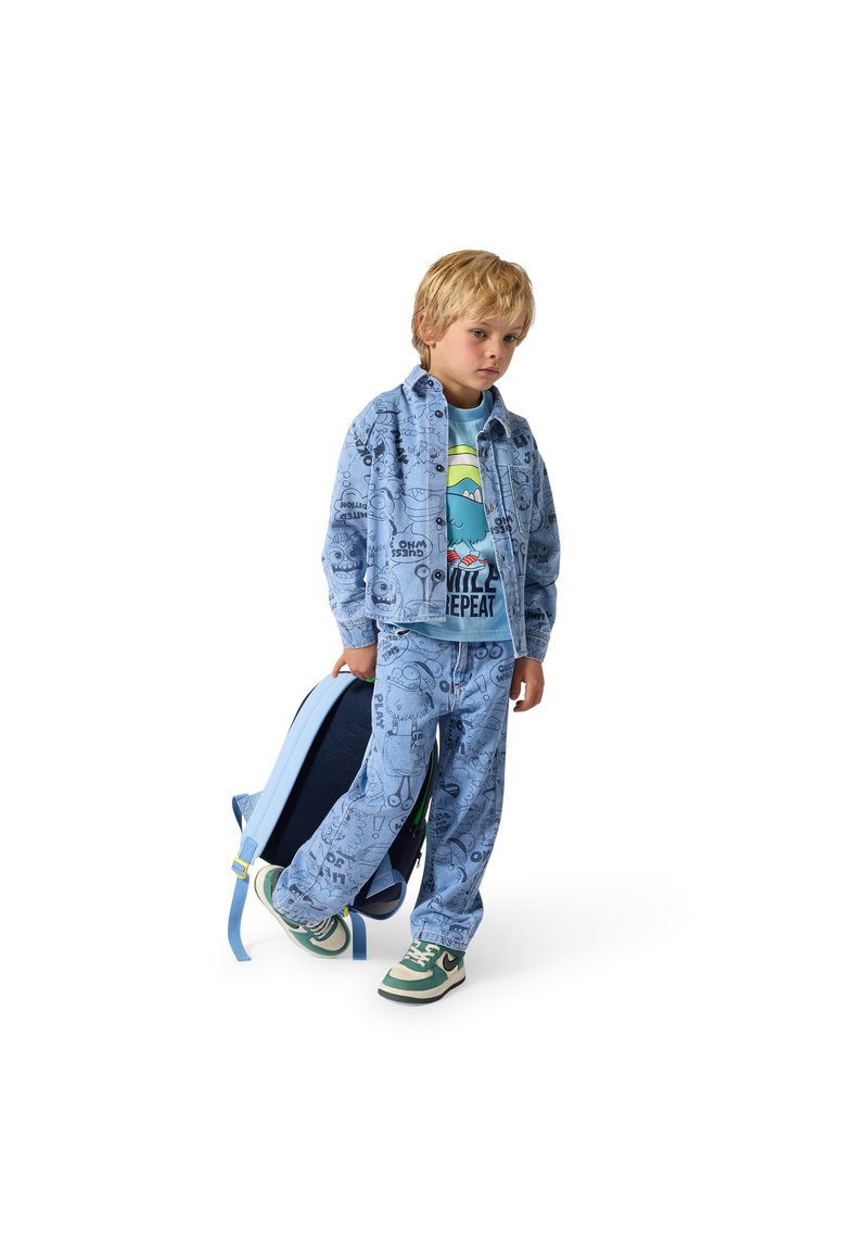 Jeune garçon portant une veste et un pantalon bleu à motifs avec un t-shirt dessin animé, tenant un sac à dos bleu, debout sur un fond blanc.