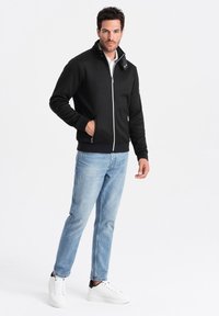 Giacca nera con zip e colletto alto, polsini a coste, tasche laterali e zip bianca. Abbinata a jeans azzurri e sneakers bianche.