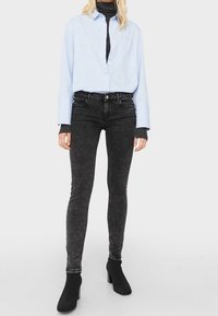 Camisa de botón azul claro, jeans negros ajustados y botas negras de tobillo. La camisa tiene un bolsillo y los jeans tienen una textura desgastada.