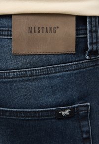 Vaqueros de mezclilla azul oscuro con un parche de cuero marrón etiquetado "MUSTANG" y una pequeña etiqueta negra con un logo de caballo en el bolsillo trasero.