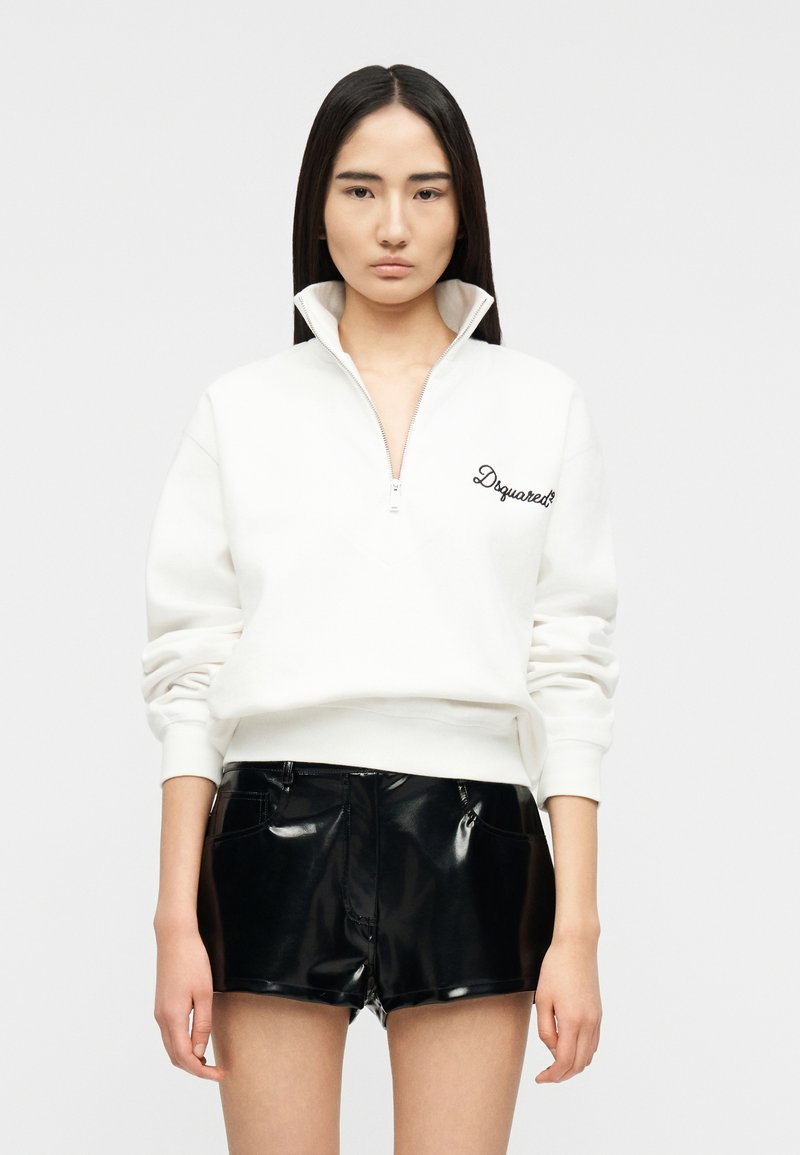 Femme aux longs cheveux bruns portant un sweat-shirt blanc à col zippé jusqu'au quart avec une écriture noire et un short noir brillant, debout devant un fond uni.