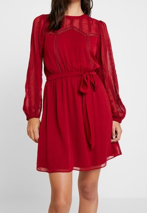 Kobieta w czerwonej sukience do kolan z przezroczystymi długimi rękawami, ozdobioną koronką i paskiem wiązanym w talii na jednolitym tle.