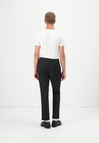 Homme portant un t-shirt blanc, un pantalon gris foncé sur mesure, des chaussettes blanches et des chaussures de ville noires, se tenant face à un fond blanc uni.