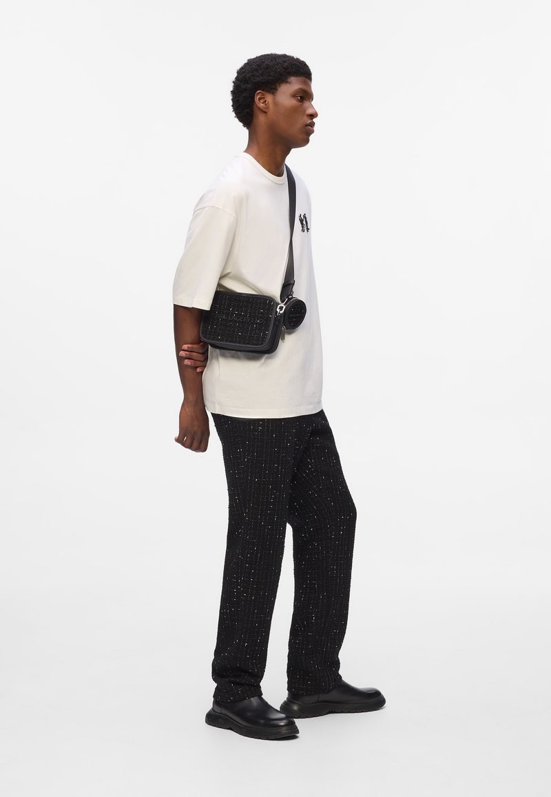 Black crossbody bag with textured surface and logo, worn with loose white t-shirt and black patterned trousers. Model stands in profile.