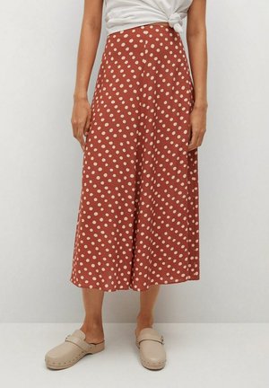 Femme portant une jupe midi couleur rouille à pois blancs, des sabots beiges et un haut blanc noué, debout devant un fond uni.