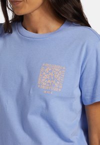 Camiseta de algodón azul claro con escote redondo, que presenta un estampado gráfico de flores color durazno cerca del área del pecho izquierdo.