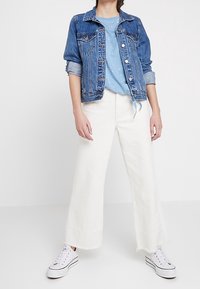 Jeansjacka med knappstängning, ljusblå skjorta med vita prickar, vida vita byxor med fransade sömmar, tillsammans med vita sneakers.