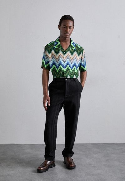 Chemise à manches courtes avec un motif en zigzag dans des tons de vert, bleu et marron ; associée à un pantalon noir texturé et des chaussures marron.