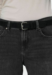 Ceinture en cuir noir avec boucle argentée portée avec un jean en denim gris foncé et une chemise blanche rentrée.