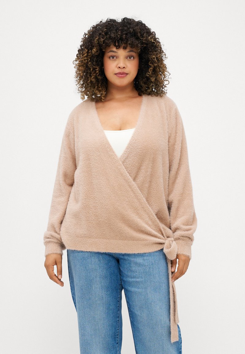 Μπεζ πλεκτό πουλόβερ με μακριά μανίκια, φτιαγμένο από μαλακό, υφασμάτινο υλικό, που διαθέτει λεπτομέρεια με κορδόνι στο πλάι και σχέδιο βαθούλωτη λαιμόκοψη. Συνδυάζεται με μπλε τζιν.