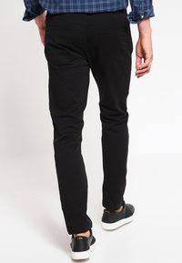 Pantalon noir en coton coupe slim avec deux poches arrière, associé à des baskets noires à semelles blanches. La personne est vue de dos.