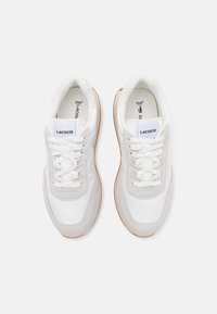 Pár bílých tenisek Lacoste s béžovými semišovými detaily, bílými tkaničkami a gumovými podrážkami, pohled zhora.