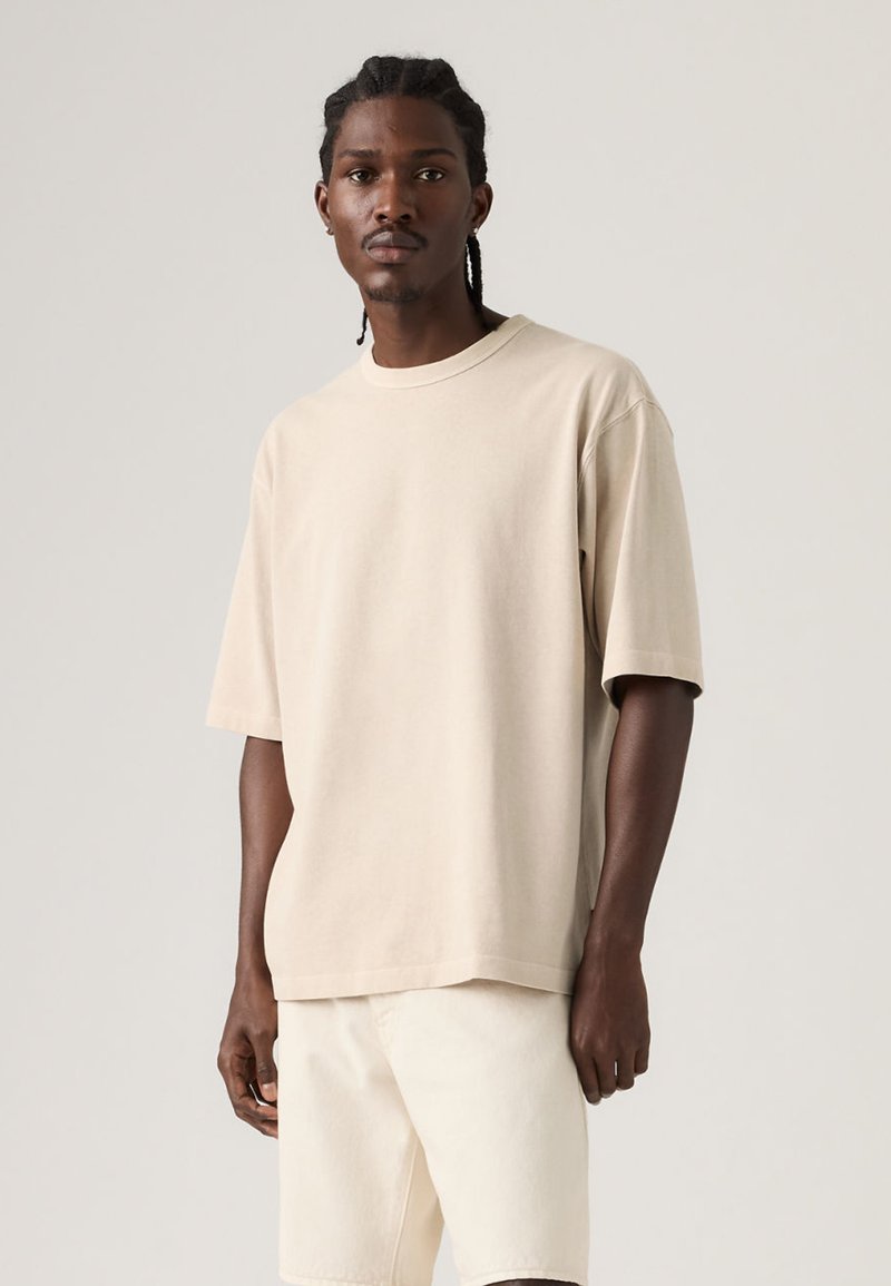 Άνδρας με πλεξουδάκια, φορώντας μπεζ πουκάμισο με κοντά μανίκια σε μεγάλο μέγεθος και κρεμ σορτς, σε απλό φωτεινό φόντο.