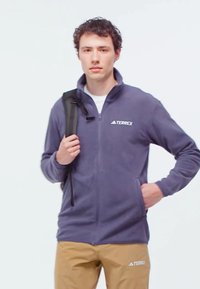 Navy fleece zip-up jacket with high collar, featuring a TERREX logo. Paired with tan pants, model holds a backpack strap in one hand.