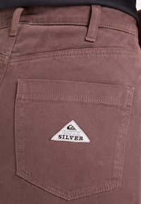 Ruskeat puuvillahousut sisältävät takataskun, jossa on kolmiomainen valkoinen etiketti, jossa lukee "QUIKSILVER" mustalla tekstillä. Huomaamaton rakenne näkyy.