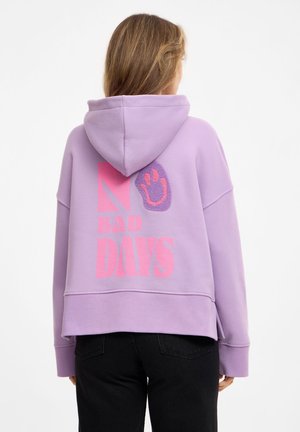Woman wearing a lavender hoodie with pink text "NO BAD DAYS" and a purple smiley face design on the back, paired with black pants.