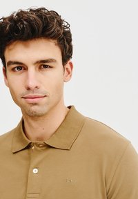 Brązowa koszulka polo z klasycznym kołnierzykiem, z dwiema guzikami i subtelnym haftowanym logo na piersi. Gładka, bawełniana tkanina o miękkiej teksturze.