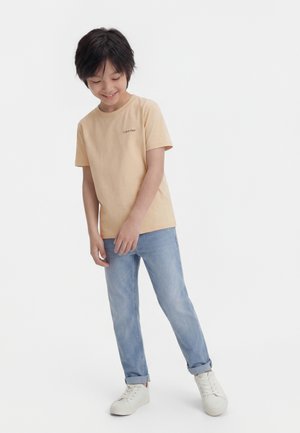 Usmievajúce sa dieťa s krátkymi čiernymi vlasmi, oblečené v béžovom tričku Calvin Klein, svetlomodrých džínsových nohaviciach vyhrnutých hore a bielych teniskách, stojace na bielom pozadí.