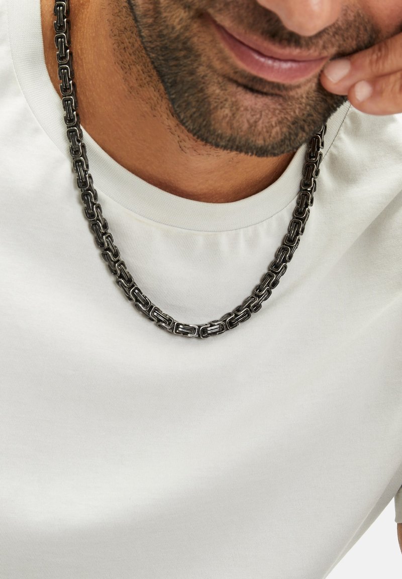 Zilverkleurige kettinghalsketting van onderling vergrendelde rechthoekige schakels, liggend op een lichtgekleurde shirt met een gladde textuur.