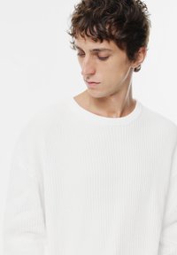 Junger Mann mit kurzwelligen braunen Haaren, der einen weißen Rippenpullover trägt und nach unten und zur Seite schaut, vor einem schlichten weißen Hintergrund.
