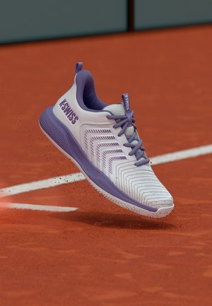 Bílo-fialová tenisová bota K-Swiss ve vzduchu nad antukovým kurtem, kolem podrážky se rozptylují částice červené antuky.