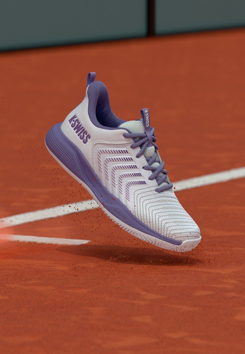 Λευκό και μωβ παπούτσι τένις K-Swiss στον αέρα πάνω από γήπεδο με χωμάτινη επιφάνεια, με κόκκινους κόκκους πηλού να σκορπίζονται γύρω από την σόλα.