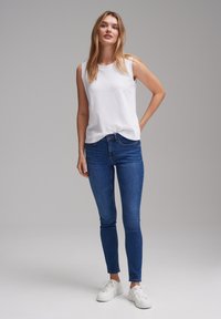 White sleeveless top, blue skinny jeans, white sneakers; casual outfit on a model with long hair, standing against a plain background.